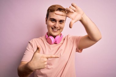 Genç, kızıl saçlı, müzik dinleyen, pembe arka planda kulaklık takan, gülümseyen, elleri ve parmakları mutlu yüzlü, çerçeveli genç bir adam. Yaratıcılık ve fotoğrafçılık kavramı.