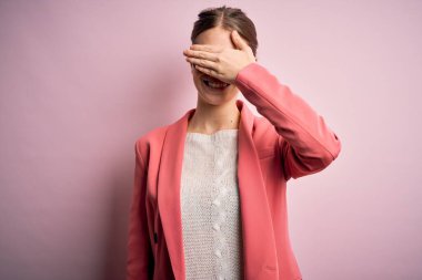 İzole edilmiş pembe arka planda ceket ve gözlük takan güzel, kızıl saçlı bir kadın. Gülümsüyor ve gülümsüyor. Yüzünde şaşkınlık için gözleri örtüyor. Kör kavram.