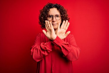 Orta yaşlı, kıvırcık saçlı, rahat gömlekli ve kırmızı arka planda gözlüklü güzel bir kadın. Reddedilmiş yüz ifadesi.