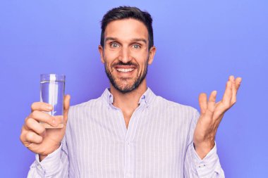 Genç yakışıklı adam, izole edilmiş mor arka planda serinlemek için bir bardak su içiyor başarıyı mutlu bir gülümsemeyle ve kazanan yüz ifadesiyle el kaldırarak kutluyor.