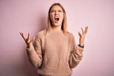 Pembe izole edilmiş arka plan üzerine kışlık yün kazak giyen genç, güzel sarışın kadın sinirli bir şekilde bağırıyor ve agresif bir ifade ve kolla bağırıyor. Hayal kırıklığı konsepti.