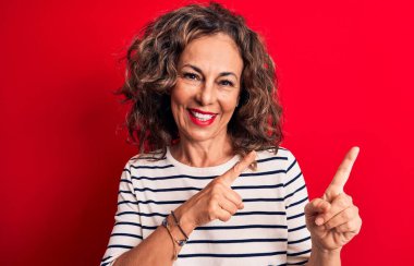 Orta yaşlı güzel esmer kadın çizgili tişört giyiyor kırmızı arka planda duruyor gülümsüyor ve kameraya bakıyor iki eliyle ve parmaklarıyla yüzünü gösteriyor..