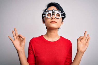 Optometri gözlüğü takan genç güzel Asyalı kız izole edilmiş beyaz arka planda duruyor rahatla ve gözleri kapalı gülümse parmaklarıyla meditasyon hareketi yapıyor. Yoga konsepti.