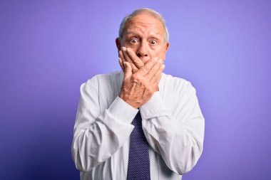 Gri saçlı, son sınıf iş adamı, mor izole arka planda dikiliyor. Ağzını yanlışlıkla ellerle kapatıyor. Gizli konsept.