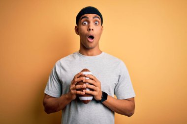 Genç, yakışıklı, Afrikalı Amerikalı bir adam ragbi topunu izole edilmiş sarı arka planda tutuyor. Şok olmuş, şaşırmış ve korku ifadesinden korkuyor.