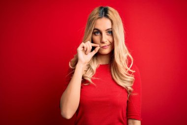Genç, güzel, sarışın, günlük tişört giyen, izole edilmiş kırmızı arka plan ağzının üzerinde duran dudakları parmaklarıyla kapanan bir kadın. Gizli ve sessiz, tabu konuşması