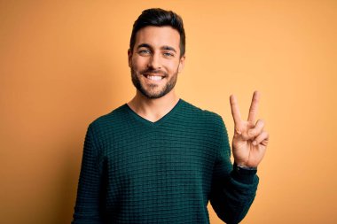 Sakallı, sıradan bir süveter giyen, sarı arka planda dikilen, kameraya gülümseyen ve zafer işareti yapan yakışıklı genç bir adam. İki numara..