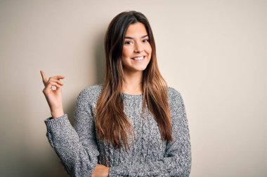 İzole edilmiş beyaz arka plan üzerinde duran sıradan bir süveter giyen genç ve güzel bir kız yüzünde kocaman bir gülümsemeyle kameraya el ve parmak ile bakıyor..