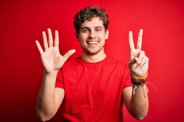 Kıvırcık saçlı, kırmızı arka planda sıradan tişörtler giyen genç, sarışın, kendine güvenen ve mutlu bir şekilde 7 numarayı işaret eden adam..