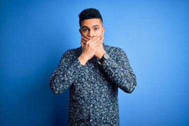 İzole edilmiş mavi arka plan üzerinde duran sıradan tişört giyen genç yakışıklı adam yanlışlıkla elleri ile ağzını kapatırken şok oldu. Gizli konsept.