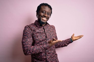 Genç, yakışıklı, Afro-Amerikan bir adam, günlük gömleğini giymiş pembe arka planda duruyor ve gülümseyerek içeri girmeye davet ediyor.