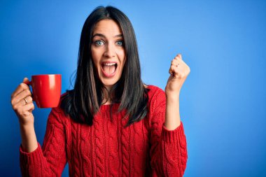Mavi gözlü genç bir kadın izole edilmiş arka planda dikilmiş bir fincan kahve içiyor gururla haykırıyor, zaferi ve başarıyı kutluyor çok heyecanlı, coşkulu bir duygu.