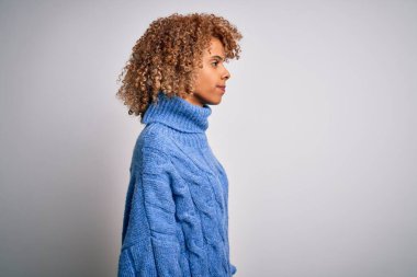 Beyaz arka plan üzerine balıkçı yaka kazak giyen genç ve güzel Afro-Amerikan kadın profilini rahatlatmak için doğal bir yüz ifadesi ve kendine güvenen bir gülümseme takınıyor..