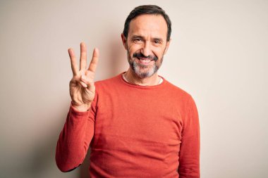 Orta yaşlı, turuncu süveter giyen, izole edilmiş beyaz arka plan üzerinde duran, üç numarayı işaret eden, kendinden emin ve mutlu gülümseyen, orta yaşlı bir adam..
