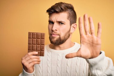 Sarışın, sakallı ve mavi gözlü bir genç. Sarı arka planda çikolata çubuğu tutuyor. Eli açık.