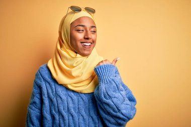 Sarı başörtüsü ve güneş gözlüğü takmış, gülümseyen, gülümseyen ve baş parmağıyla yüzünü gösteren Afro-Amerikan öğrenci kadın..