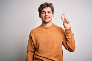 Sarışın, kıvırcık saçlı, beyaz arka planda sıradan süveter giyen, parmaklarını zafer işareti yaparken gösteren kameraya gülümseyen genç, yakışıklı bir adam. İki numara..