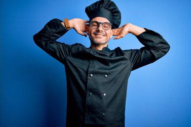 Genç, yakışıklı aşçı üniforması ve şapkası olan izole edilmiş mavi arka planda gevşeyen ve esneyen, kolları ve elleri başının arkasında ve boynunun arkasında mutlu bir şekilde gülümseyen bir aşçı.