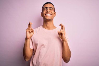 Yakışıklı Afro-Amerikalı adam pembe arka plan üzerinde günlük tişört ve gözlük takıyor. Umut ve gözler kapalı gülümseyerek işaret ediyor. Şans ve batıl inanç..