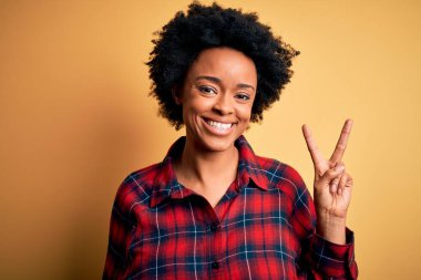 Kıvırcık saçlı, rahat gömlekli, kameraya gülümseyen ve zafer işareti yapan güzel Afro-Amerikan kadın. İki numara..