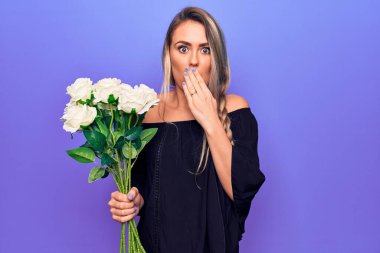Genç, güzel, sarışın bir kadın, bir buket çiçekle izole edilmiş mor arka planda, ağzı elle kaplayan, şok olmuş ve hata yapmaktan korkan bir kadın. Şaşırtıcı ifade.