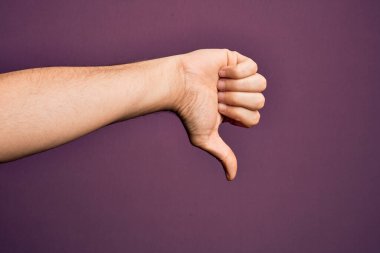 İzole edilmiş mor arka plan üzerinde parmaklarını gösteren beyaz bir gencin eli. Baş parmak reddetme hareketi, onaylanmama hareketi ve olumsuz işaret yapıyor.