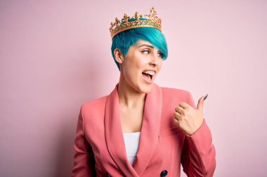 Mavi saçlı, pembe izole edilmiş arka planda kraliçe tacı takan genç bir iş kadını. Gülümsüyor ve başparmağıyla yana bakıyor..