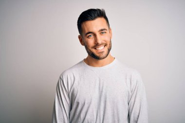 Yüzünde mutlu ve havalı bir gülümsemeyle izole edilmiş beyaz arka plan üzerinde duran sıradan tişört giyen genç yakışıklı bir adam. Şanslı kişi..