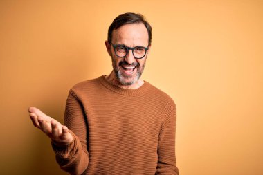 Orta yaşlı, beyaz, kahverengi süveter ve gözlük takan, izole edilmiş sarı arka planda neşeli gülümseyen, dost canlısı, olumlu ve kendinden emin bir şekilde selamlayan bir adam.