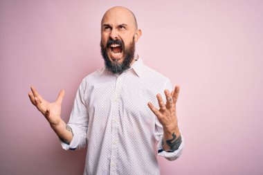 Soyutlanmış pembe arka planda zarif gömlek giyen sakallı yakışıklı kel adam deli gibi bağırıyor ve agresif bir ifadeyle bağırıyor ve kollarını kaldırıyor. Hayal kırıklığı konsepti.