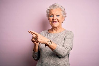 Son sınıf güzeli, rahat tişört giyen, pembe arka planda duran, gülümseyen ve kameraya bakıp iki eliyle ve parmaklarıyla kenarı gösteren bir kadın..