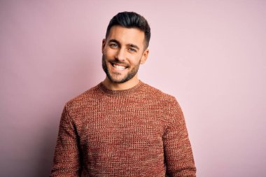 Yüzünde mutlu ve havalı bir gülümsemeyle pembe bir arka plan üzerinde duran sıradan bir süveter giyen genç yakışıklı bir adam. Şanslı kişi..