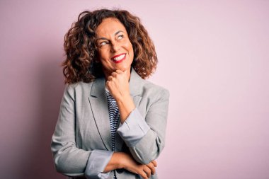 Orta yaşlı, güzel bir iş kadını, soyutlanmış pembe arka planda zarif bir ceket giyiyor, elleri çenesinde soru sorarken, düşünceli bir ifade takınıyor. Düşünceli bir yüzle gülümsüyor. Şüphe kavramı.