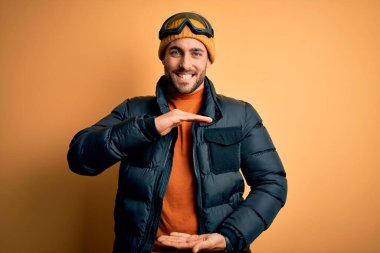 Kar giysileri ve kayak gözlüğü takan sakallı genç yakışıklı kayakçı. Büyük ve büyük beden işareti gösteren el kol hareketleri yapıyor. Ölçüm sembolü. Kameraya bakıp gülümsüyor. Ölçüm konsepti.