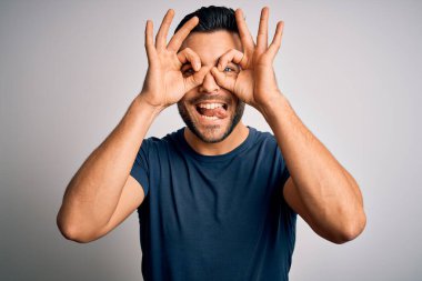 İzole edilmiş beyaz arka plan üzerinde duran sıradan tişört giyen genç yakışıklı bir adam dürbün gibi dil çıkartıyor, gözler parmaklarından bakıyor. Çılgın ifade.