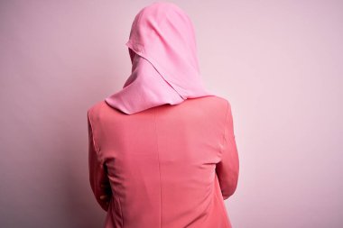 İslami başörtüsü takan genç ve güzel bir kız soyutlanmış pembe arka planda dikilip kollarını kavuşturup geriye bakıyor.