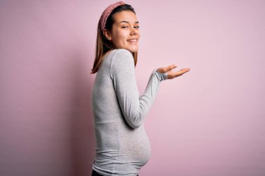 Genç, güzel bir kız hamile, pembe arka plan üzerinde bebek bekliyor. Elleri açık, fotokopi alanını gösteren avuç içleri açık, gülümseyerek mutlu bir şekilde gülümseyen bir ilan veriyor.