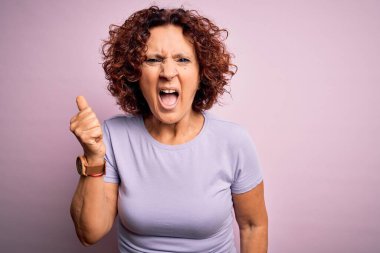 Orta yaşlı güzel kıvırcık saçlı kadın pembe arka plan üzerine gündelik tişört giyip öfkeyle bağırırken kızgın ve kızgın bir şekilde yumruğunu kaldırıyor. Öfke ve agresif kavram.