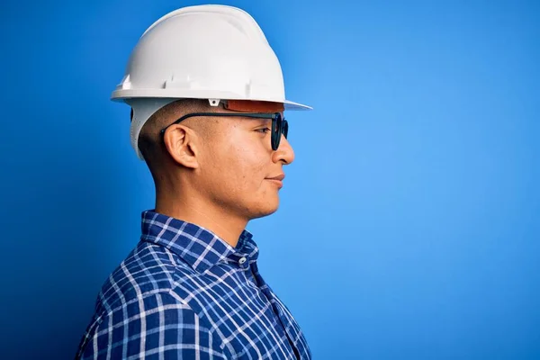 Young handsome engineer latin man wearing safety helmet over isolated blue background looking to side, relax profile pose with natural face with confident smile.