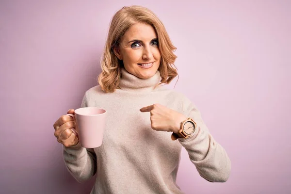 中年美丽的金发女人在与世隔绝的背景下喝着粉红色的咖啡 满脸惊讶地指着自己 — 图库照片