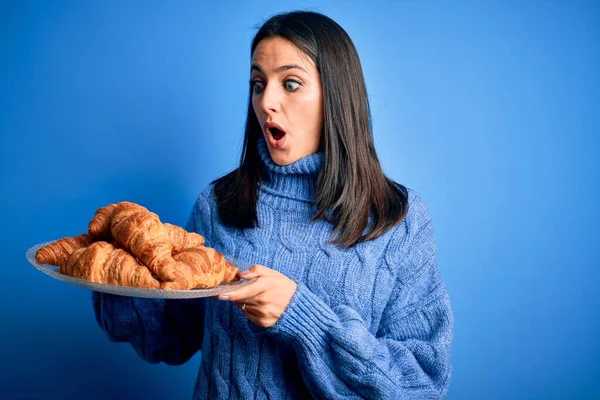 青い目の若い女性が甘いですクロワッサンを保持します朝食のために隔離された背景驚きの顔でショックで怖がっています 恐れと恐怖の表現で興奮 — ストック写真