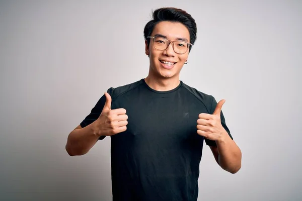Young handsome chinese man wearing black t-shirt and glasses over white background success sign doing positive gesture with hand, thumbs up smiling and happy. Cheerful expression and winner gesture.