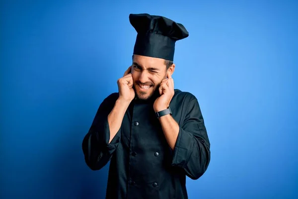 年轻英俊的厨师 留着胡子 身穿炊具制服 头戴蓝色背景帽 用手指捂住耳朵 对嘈杂的音乐感到恼怒 聋人概念 — 图库照片
