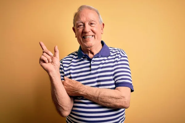 Grey haired senior man wearing casual navy striped t-shirt standing over yellow background with a big smile on face, pointing with hand and finger to the side looking at the camera.
