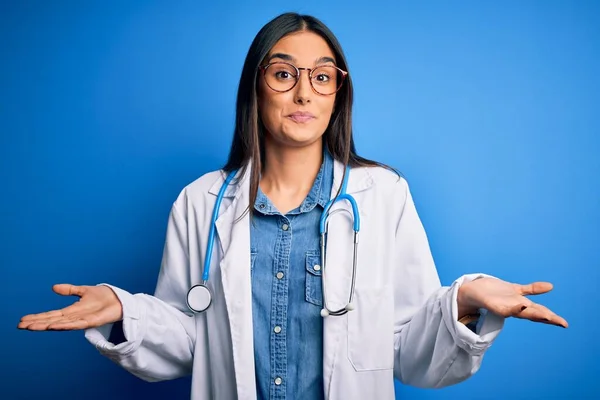 Young beautiful doctor woman wearing stethoscope and glasses over blue background clueless and confused expression with arms and hands raised. Doubt concept.