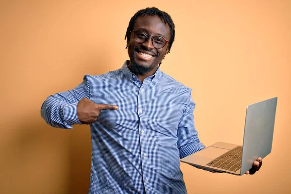 若いアフリカ系アメリカ人の労働者の男作業ノートパソコンを使用して黄色の背景に立って驚きの顔を指差す指彼自身 — ストック写真