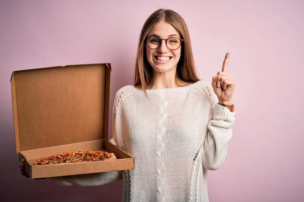 美しいです赤毛女性保持配達ボックスとともにイタリアの新鮮なピザ以上ピンクの背景驚きでアイデアや質問指差し指で幸せな顔 ナンバーワン — ストック写真