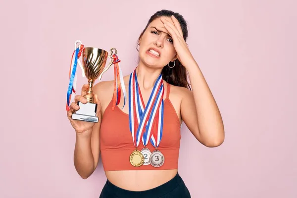 Young beautiful fitness winner athlete woman wearing sport medals and trophy stressed with hand on head, shocked with shame and surprise face, angry and frustrated. Fear and upset for mistake.