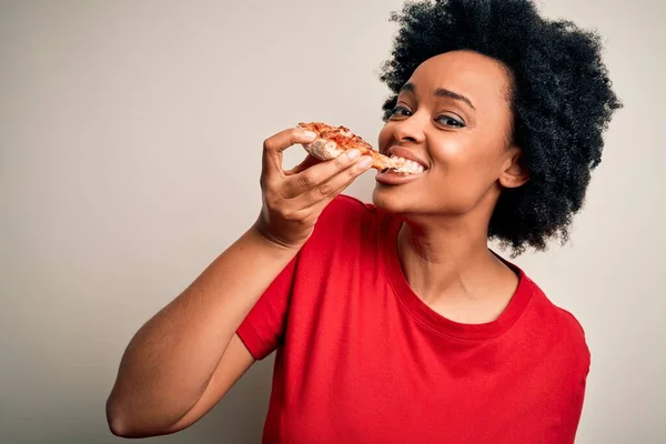 Young beautiful african american afro woman smiling happy and confident. Standing with smile on face eating delicious slice of italian pizza over isolated white background