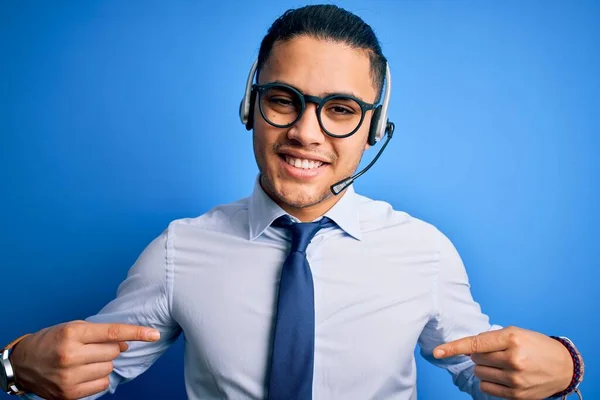 Young brazilian call center agent man wearing glasses and tie working using headset looking confident with smile on face, pointing oneself with fingers proud and happy.